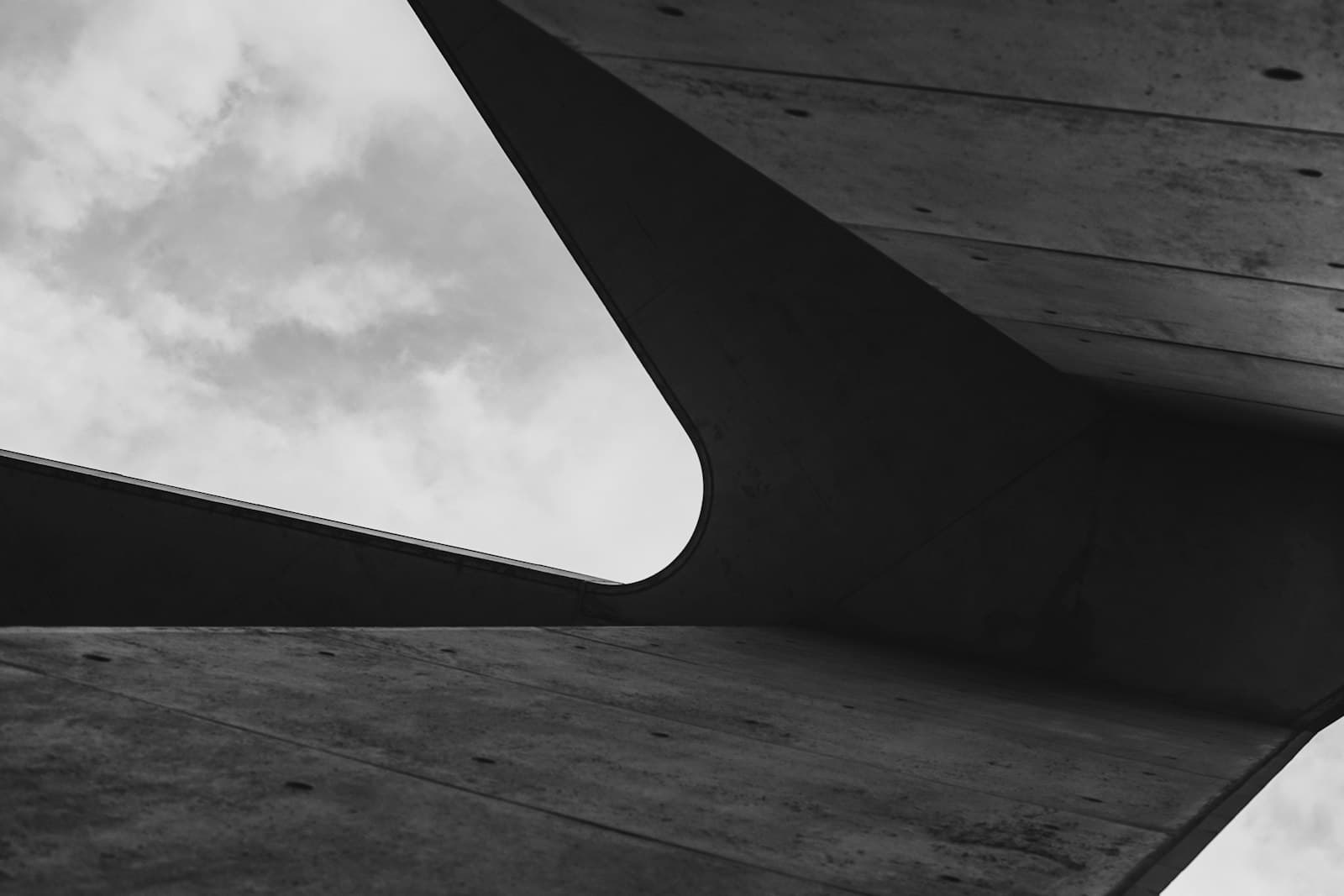 Underside of a brutalist concrete overpass, geometric facets converging against a gray sky.