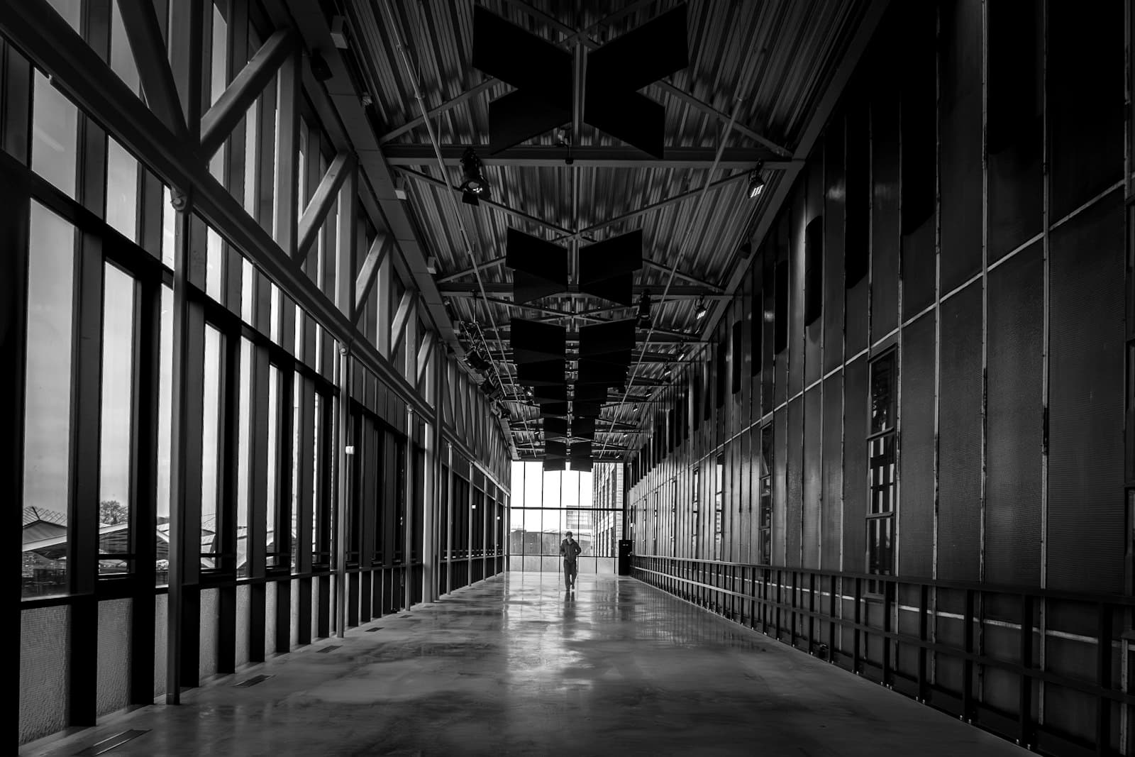 Pedestrian walking the length of a long modernist corridor, framed by parallel girders and glass.