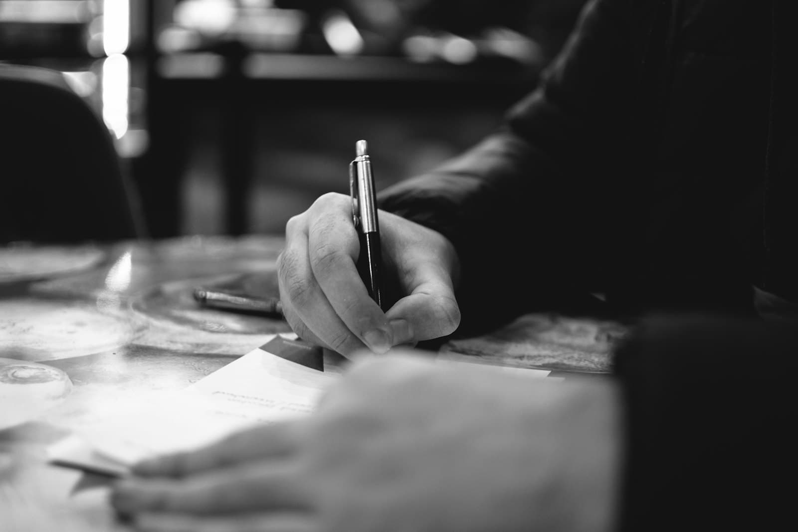 A hand holding a pen above a printed document on a desk, second hand resting on the page.