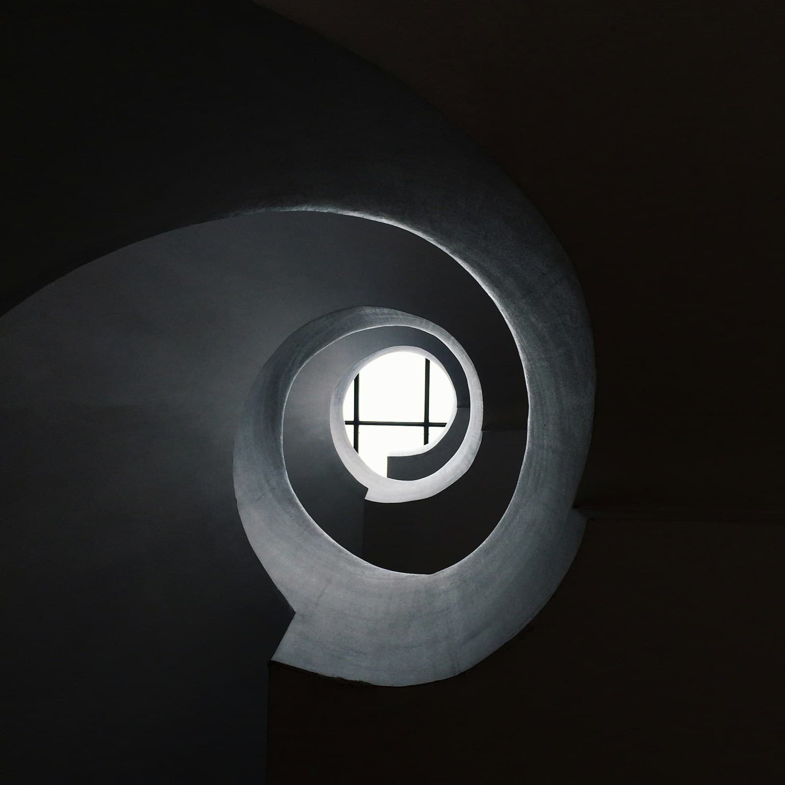 Bottom view of a smooth concrete spiral stair curving around a small circular skylight, the left half falling into deep shadow.
